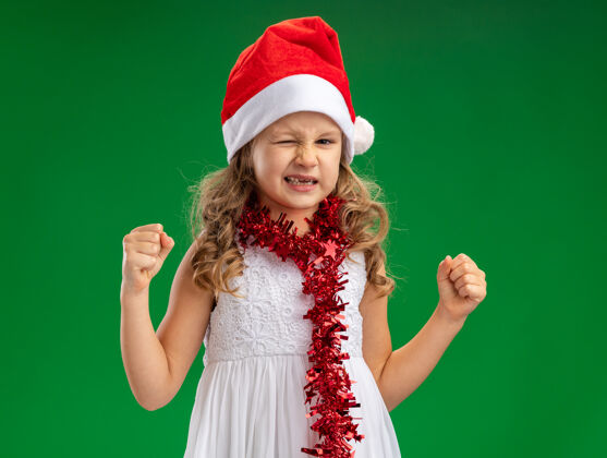 兴奋兴奋地眨着眼睛的小女孩戴着圣诞帽 脖子上戴着花环 在绿色背景上显示“是”的手势眨眼圣诞秀