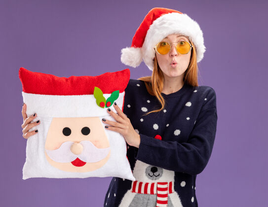 关心关心的年轻漂亮女孩穿着圣诞毛衣和帽子戴着眼镜拿着圣诞枕头隔离在紫色的背景上女孩毛衣年轻