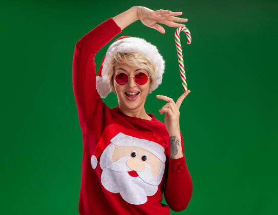 金发快乐的金发女郎戴着圣诞帽和圣诞老人的圣诞毛衣戴着眼镜看着相机拿着圣诞糖果手杖靠近头部隔离在绿色背景上空间圣诞拿着