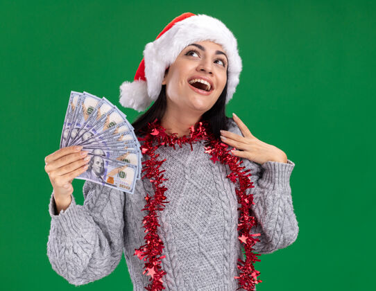 绿色令人印象深刻的年轻白人女孩戴着圣诞帽 脖子上戴着金箔花环 手里拿着钱 抚摸着肩膀 在绿色的背景下孤零零地抬头仰望金属片圣诞快乐触摸