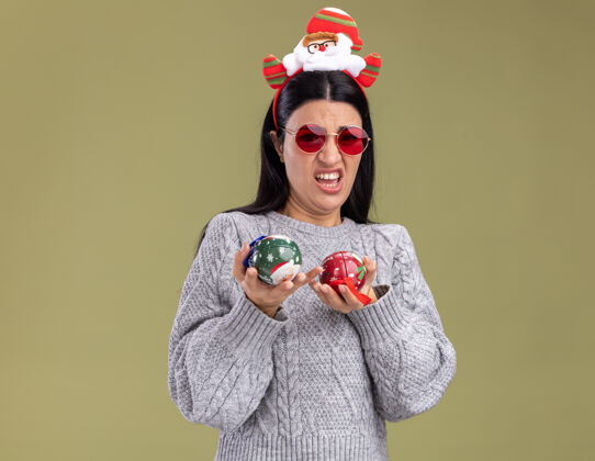 绿色恼怒的年轻白人女孩戴着圣诞老人的头带 戴着眼镜 拿着圣诞饰品 看着橄榄绿背景上的相机举行年轻圣诞
