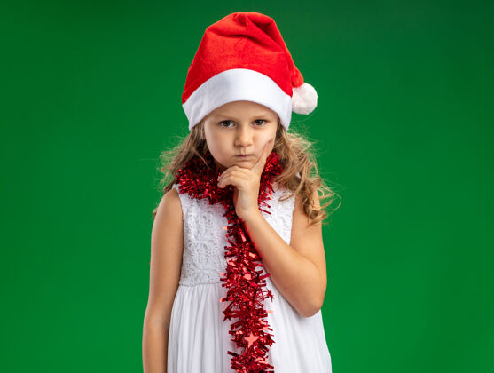 下巴皱着眉头的小女孩戴着圣诞帽 脖子上戴着花环 手放在下巴上 绿色的背景上孤立着绿色脖子手