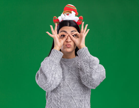 年轻令人印象深刻的年轻白人女孩戴着圣诞老人的头带看着相机做看手势用手作为望远镜隔离在绿色背景上女孩头带手
