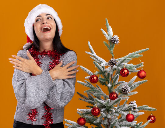 向上令人印象深刻的年轻白人女孩戴着圣诞帽 脖子上戴着金箔花环 站在装饰过的圣诞树旁 双手交叉放在胳膊上 在橙色背景下孤立地抬头看手臂帽子十字架