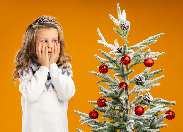 花环关心的小女孩站在圣诞树旁 戴着头饰 脖子上戴着花环 两手被隔离在橙色的背景上手小橘子