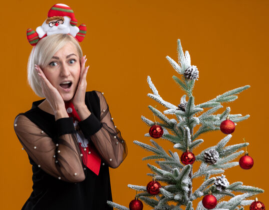 脸兴奋的中年金发女人戴着圣诞老人的头带和领带站在装饰过的圣诞树旁 手放在脸上看着橙色背景上的相机金发中年站着