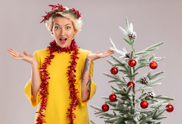 花圈令人印象深刻的年轻金发女子头戴圣诞花环 脖子上戴着金属丝花环站在装饰过的圣诞树旁 看着镜头 在白色背景上孤立着空空的双手空金发金属片
