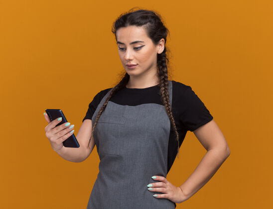人印象深刻的年轻女理发师在制服举行 并在电话看把手放在臀部隔离在橙色墙上感觉拿着女性