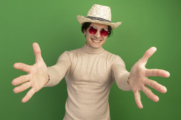 帽子面带微笑的年轻帅哥戴着帽子 戴着眼镜 在绿色的墙前伸出双手帅气衣服眼镜