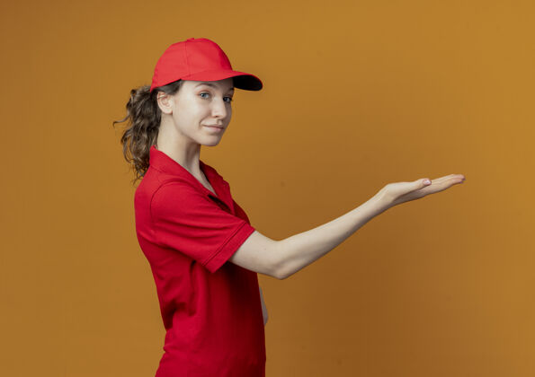 个人资料穿着红色制服 戴着帽子的年轻漂亮的送货女孩站在侧视图中 用手直指 看着隔离在橙色背景上的相机 还有复印空间请帽子背景