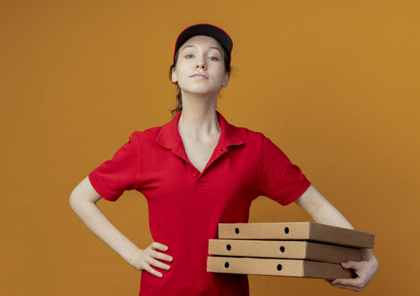 手自信的年轻漂亮的送货女孩 穿着红色制服 戴着帽子 手里拿着披萨包 手放在腰上 看着橙色背景上孤立的摄像机送货披萨帽子