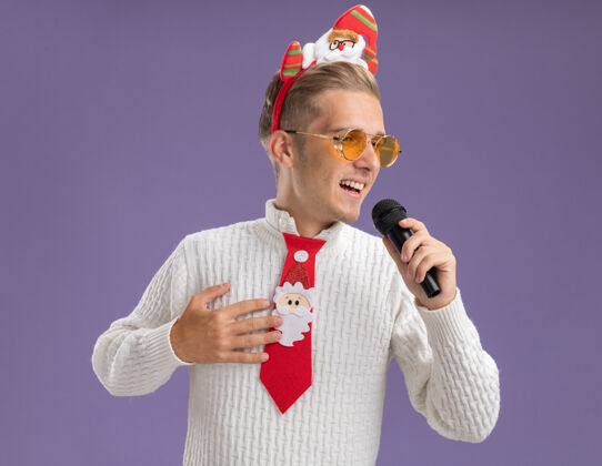 圣诞快乐快乐的年轻帅哥戴着圣诞老人的头带和领带戴着眼镜拿着麦克风手放在胸前看着边唱歌紫色背景上孤立快乐圣诞老人抱着