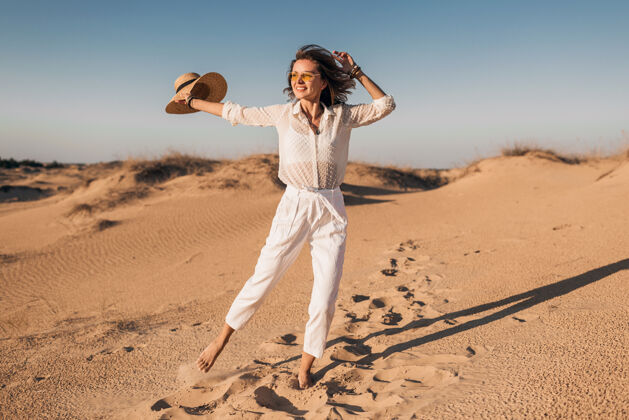 体验时髦的微笑美丽的快乐女人穿着白色的衣服在沙漠中奔跑和跳跃在日落时戴着草帽快乐女人旅游