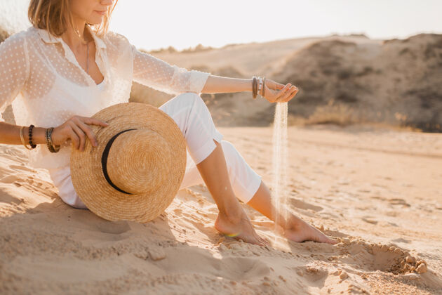 沙滩落日时分 沙漠中穿着白衣 头戴草帽的时尚美女用沙子将双手合拢人裤子衣服