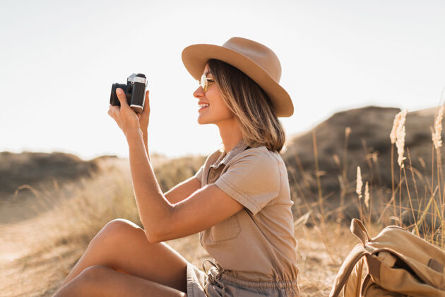 旅游沙漠中穿着卡其色长裙的迷人时尚年轻女子 在非洲旅行 戴着帽子和背包 用老式相机拍照 探索大自然 阳光明媚的天气时尚配件沙丘