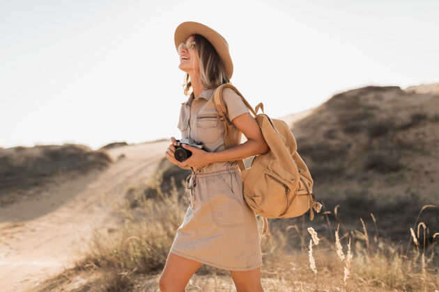 沙漠沙漠中穿着卡其色长裙的迷人时尚年轻女子 在非洲旅行 戴着帽子和背包 用老式相机拍照游客配件背包