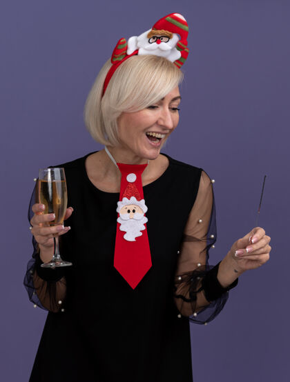 领带快乐的中年金发女人戴着圣诞老人的头带 打着领带 手里拿着节日火花和香槟 看着紫色背景上孤立的火花欢乐头带金发