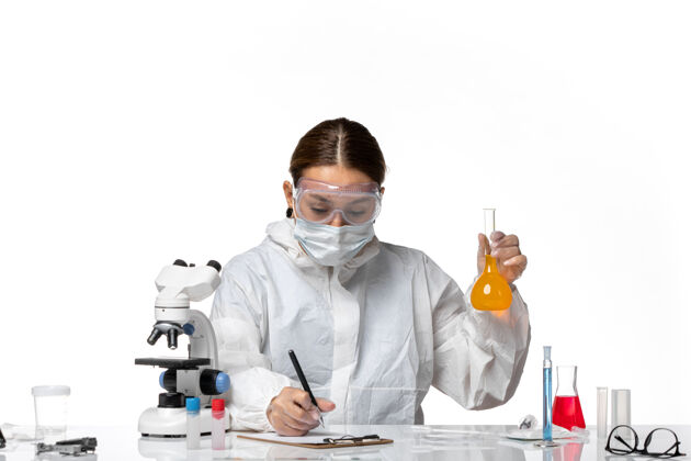 白色正面图女医生穿着特制的工作服 戴着口罩 拿着装有黄色溶液的烧瓶 白色背景上有冠状病毒大流行病毒实验室专业