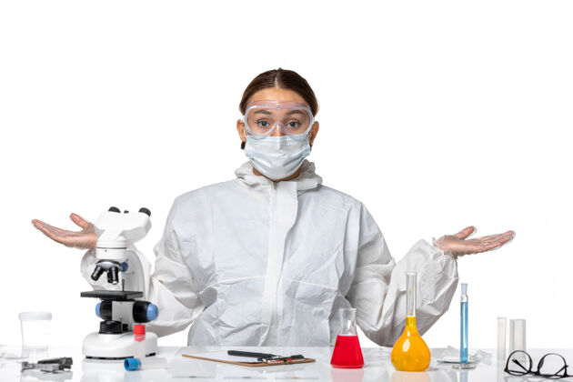 耸肩正面图：穿着防护服 戴着口罩的女医生坐在白色办公桌上解决病毒-健康大流行医疗实验服视野