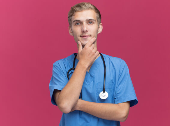 男性高兴的年轻男医生穿着医生制服和听诊器抓住下巴孤立在粉红色的墙上站表情衣服