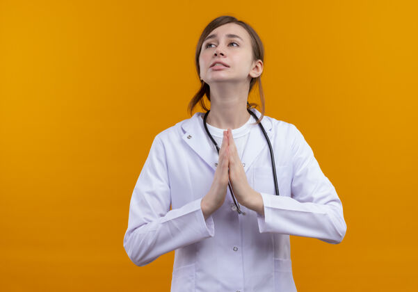 年轻焦急的年轻女医生穿着医用长袍和听诊器在隔离的橙色空间和复印空间做祈祷手势医疗祈祷医生