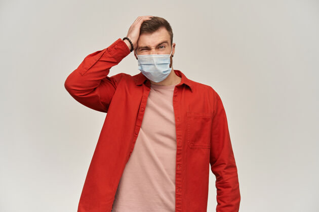 医学穿着红衬衫 脸上戴着防冠状病毒的防毒面具 满脸愁容 愁眉苦脸的年轻人 手放在头上 在白墙上头痛英俊成人人