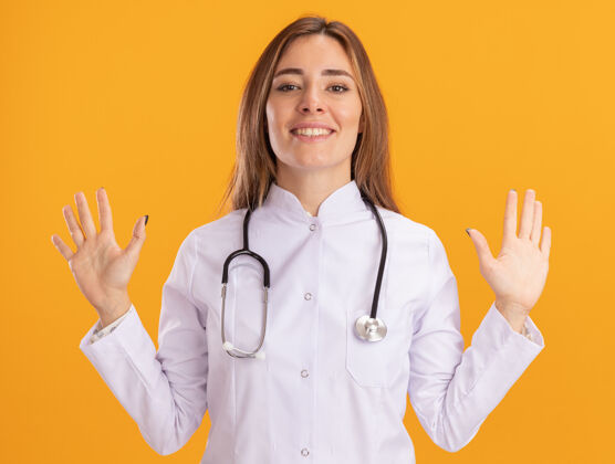表情面带微笑的年轻女医生穿着医用长袍 听诊器摊开双手隔离在黄色的墙上长袍手人