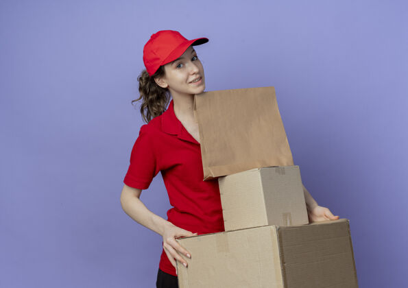 漂亮令人高兴的年轻漂亮的送货女孩穿着红色制服和帽子举行纸箱和纸包装隔离在紫色背景与复制空间女孩帽子制服