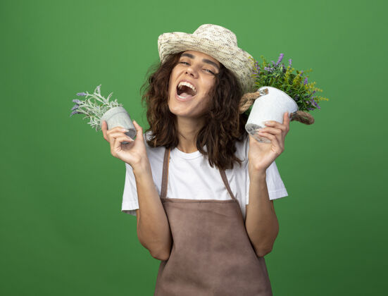 绿色兴奋的年轻女园丁穿着制服 戴着园艺帽 手里拿着隔离在绿色花盆里的花女性园艺花