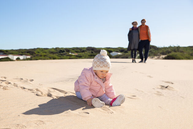 坐着可爱的宝宝穿着粉色的夹克和帽子 在户外的沙滩上玩耍衣服小为人父母