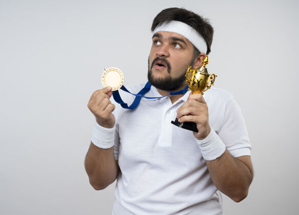 年轻惊讶的年轻人戴着头带和腕带拿着冠军杯戴着奖牌隔离在白墙上奖杯男子穿