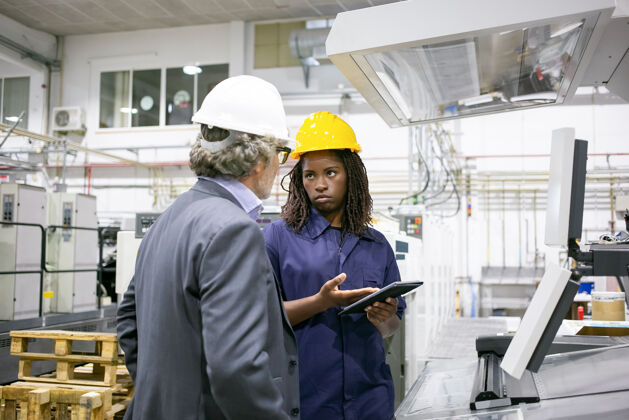 显示严肃的女工厂工人在车间的机器前和老板说话 指着平板电脑屏幕安全帽机器技术