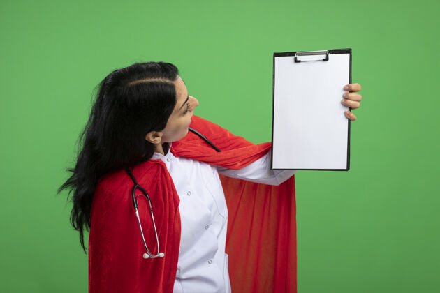 超级英雄惊讶的年轻超级英雄女孩穿着医用长袍 听诊器抬起 看着绿色的剪贴板长袍听诊器剪贴板