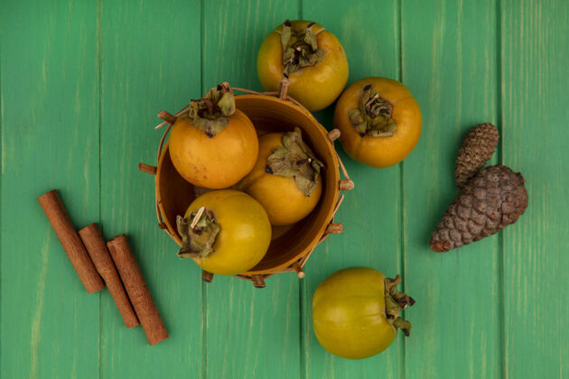 肉桂绿色木桌上放着肉桂棒的木桶上新鲜柿子水果的俯视图绿色秋天柿子
