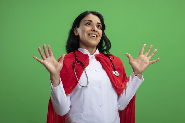 手惊讶的年轻超级英雄女孩穿着医用长袍听诊器摊开双手隔离在绿色年轻长袍超级英雄