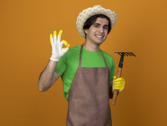 穿年轻的男园丁面带微笑 穿着制服 戴着园艺帽 戴着手套 拿着耙子 在橘子上表现出良好的姿态橙色帽子男性