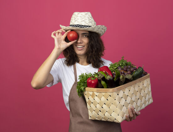 制服身着制服的年轻女园丁面带微笑 戴着园艺帽 手里拿着菜篮子 展示着西红柿孤立在粉红色上的表情粉色篮子举行