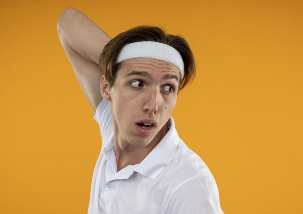 脖子年轻的运动型男人一边看一边戴着头带和腕带把手放在脖子上隔离在橙色的墙上壁板运动穿
