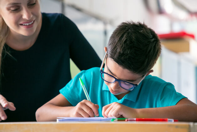 女人快乐的学校老师看着戴眼镜的男生在课堂上做作业青年知识学习