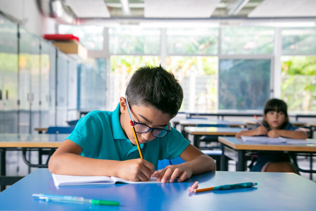 女孩一个戴眼镜的专注的小男孩坐在课桌旁 在课堂上用文案写字拿着室内室内