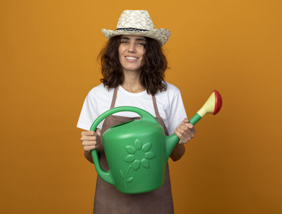 举行微笑着的年轻女园丁 穿着制服 戴着园艺帽 手里拿着浇水罐年轻人微笑园艺