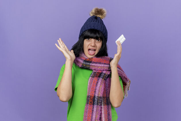 围巾印象深刻的年轻生病的女人戴着冬天的帽子和围巾拿着一包药片显示空手看前面孤立的紫色墙壁与复制空间站印象包