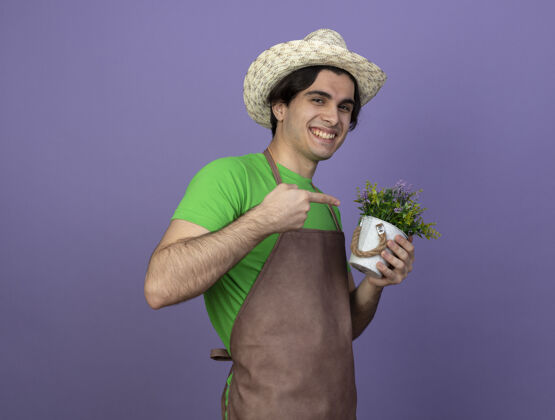 花微笑着的年轻男园丁穿着制服 戴着园艺帽 指着花盆里的花人帽子紫色
