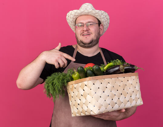 花园年轻的男园丁高兴地戴着园艺帽 指着隔离在粉红色墙上的菜篮子年轻点请