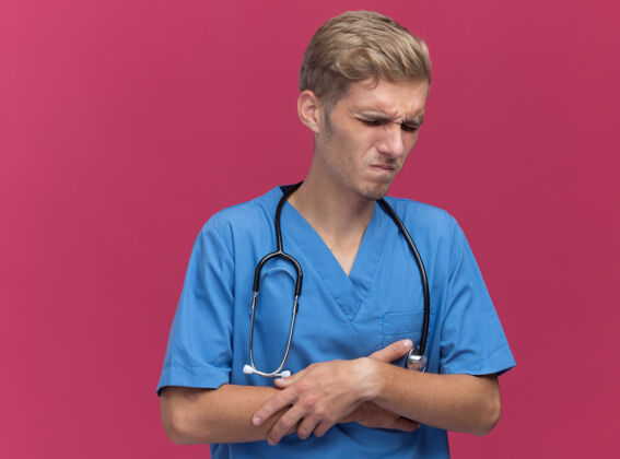 男性年轻的男医生穿着医生制服 戴着听诊器 把疼痛的胃隔离在粉红色的墙上医生抓制服