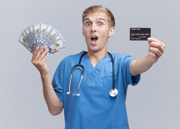 医生惊讶的年轻男医生穿着医生制服手持听诊器拿出现金和信用卡隔离在白墙上穿惊喜信用