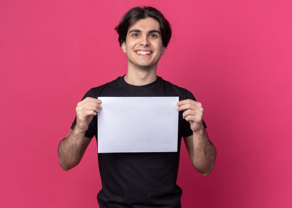 抱着面带微笑的年轻帅哥穿着黑色t恤拿着纸隔离在粉红色的墙上穿微笑小伙子