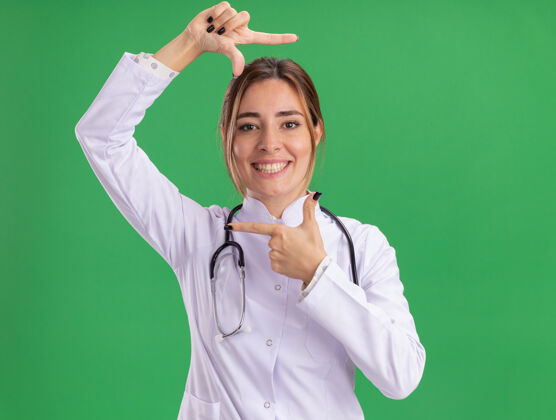 微笑微笑的年轻女医生穿着医用长袍 带着听诊器 在绿色的墙上展示着照片的姿势医生长袍女性
