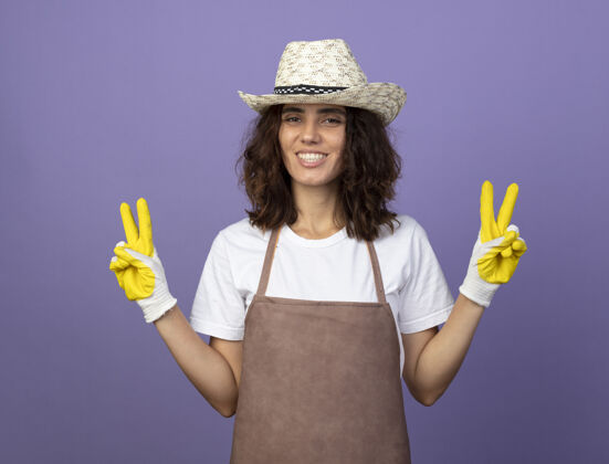 制服微笑的年轻女园丁穿着制服 戴着园艺帽和手套 显示出和平的姿态年轻紫色成人
