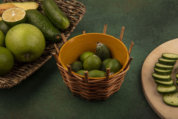 托盘俯视图：绿色背景上的柳条托盘上放着绿色苹果 鳄梨和黄瓜 桶上放着切碎的黄瓜片苹果柳条黄瓜